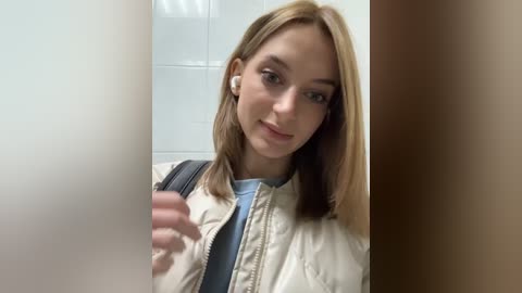 Media: Video of a young Caucasian woman with straight, shoulder-length blonde hair, wearing a white leather jacket over a light blue shirt, standing in a tiled bathroom, holding a white phone.