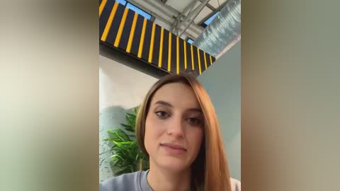 Media: Video of a young woman with long, straight brown hair, wearing a gray shirt, standing in an indoor setting with yellow and black striped ceiling panels and green plants.