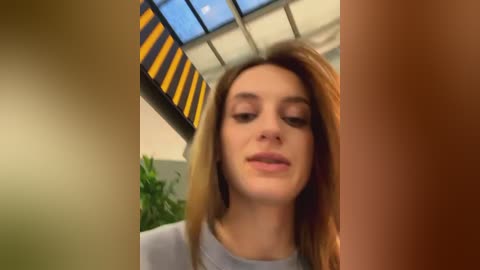 Media: Video of a young Caucasian woman with straight brown hair, fair skin, and light makeup. She's indoors near a glass ceiling with yellow and black striped panels, wearing a light-colored top.