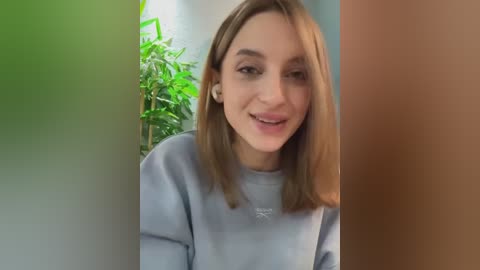 Media: Video of a smiling young woman with light skin and straight, shoulder-length brown hair, wearing a light gray sweatshirt. She stands in a room with green potted plants and a brown door in the background.