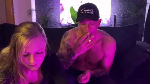 Media: Video of a tattooed, shirtless man with a cap smoking next to a blonde woman on a dark couch, lit by purple lights, in a modern living room.