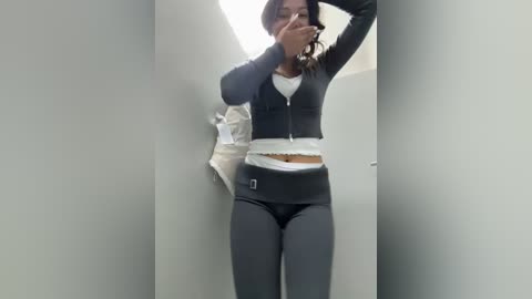 Media: A video of a woman with medium-dark skin, wearing a black zip-up hoodie, white crop top, and black leggings, posing with her hands on her head in a narrow, dimly lit hallway.