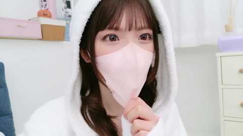 Media: Video of a young Asian woman with straight brown hair, wearing a white robe and pink face mask, holding a white tissue, in a brightly lit bathroom with white and pastel-colored decor.