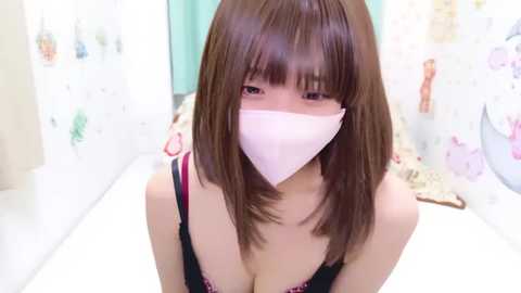 Media: A video of an Asian woman with straight brown hair and bangs, wearing a pink face mask, black bra, and light-colored shirt. Background features pastel-colored wall art and a bed.