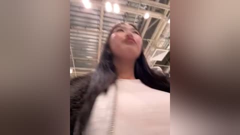 Media: A video captures a woman with long black hair and light skin, wearing a white top and black jacket, standing in an industrial warehouse with exposed beams and overhead lights.