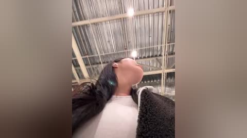 Media: Video of a person with long, dark hair lying on their back on a white bed, under a corrugated metal ceiling with visible industrial lights, wearing a white shirt and gray towel.