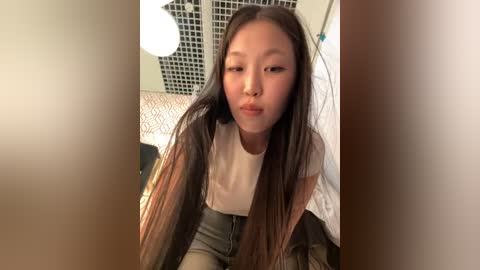 Media: Video of an East Asian woman with long, straight black hair, light skin, and a white t-shirt, making a kissy face while sitting on a bed with a white quilt and black and white geometric patterned pillow.