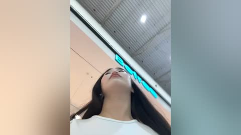 Media: Video of a young woman with long, straight dark hair and fair skin, wearing a white shirt, standing in a modern, brightly lit room with a tiled ceiling and a large digital display screen.