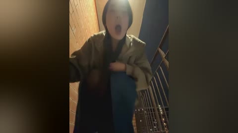 Media: Video of a young Asian woman, hooded, mouth open in shock, climbing a stairwell with brick walls, metal railings, and a blue door in the background.