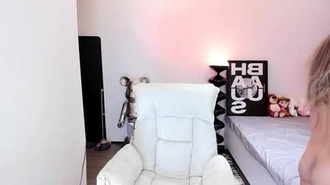 Media: Video of a minimalist bedroom with a white leather armchair, black wall art, a plush toy bear, and a pink lamp, partially visible person standing.