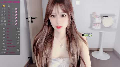 Media: A video of a young Asian woman with fair skin, long brown hair, and red lipstick, wearing a strapless white dress. She stands in a modern, minimalist room with a white table and a door in the background.