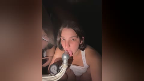 Media: Video of a young woman with medium-length dark hair, wearing a white tank top, licking the rim of a glass, with a dimly lit, dark background.