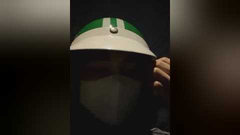 Media: Video of a person wearing a white and green hat with a black face mask, holding a black object, in a dimly lit, indistinct background.