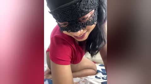 Media: Video of a woman with long black hair, wearing a black lace mask and red top, kneeling on a bed with white and blue patterned sheets, smiling.