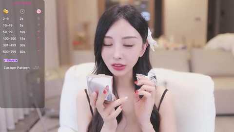 Media: A video of an East Asian woman with long black hair, wearing a white ribbon, holding a cupcake, seated indoors on a white couch.