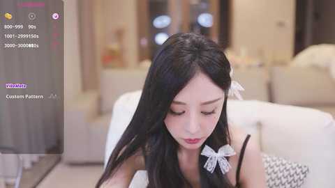 Media: Video of an Asian woman with long black hair, wearing a white bow, sitting in a modern living room with beige furniture.