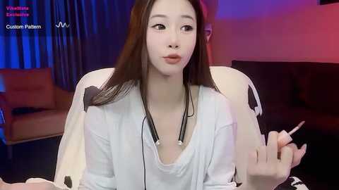 Media: Video of an Asian woman with fair skin and long black hair, wearing a loose white top, smoking a cigarette, sitting in a dimly lit room with blue and red lighting.