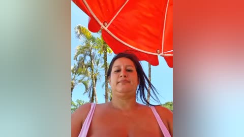 Media: Video of a woman with long black hair, wearing a pink tank top, under a red beach umbrella, with palm trees in the background on a sunny day.