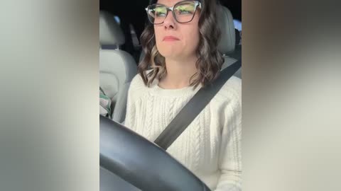 Media: Video of a woman with shoulder-length, wavy brown hair and glasses, wearing a cream-colored sweater, seated in a car, with a black seatbelt across her chest.