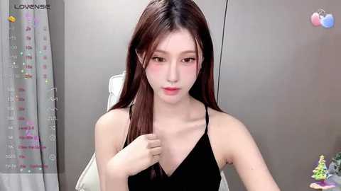 Media: Video of a young East Asian woman with fair skin, long brown hair, and a slender build, wearing a black sleeveless top, sitting in a modern room with gray walls and a colorful calendar.