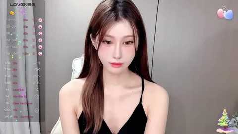 Media: A video of an East Asian woman with long, straight brown hair, fair skin, and small breasts, wearing a black spaghetti-strap top, seated indoors. Background includes a grey wall and a white curtain.