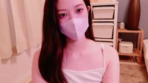 Media: Video of an Asian woman with long dark hair, wearing a white mask, white tank top, and seated indoors, surrounded by beige curtains, white shelves, and baskets.