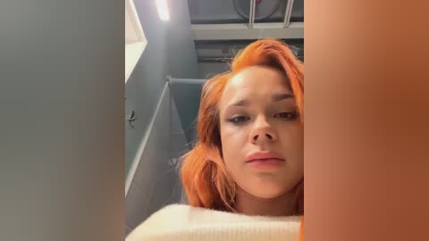 Media: A video of a young woman with striking orange hair and light skin, wearing minimal makeup, standing in a narrow, dimly lit corridor. Her expression is neutral.