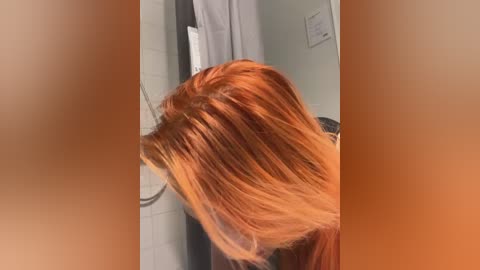 Media: Video of a woman with long, reddish-brown hair, styled in a sleek, straight bob cut, standing in a bathroom with a white-tiled wall and a partially visible showerhead.