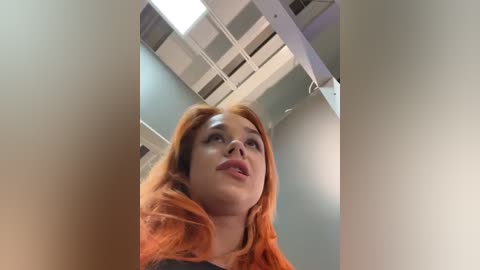 Media: Video of a young woman with long, vibrant orange hair, fair skin, and full lips, looking up. She stands in a modern, minimalist room with white ceiling tiles and a rectangular skylight.