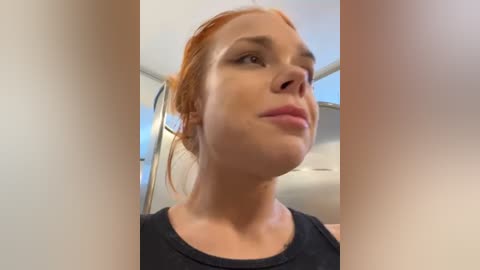 Media: Video of a Caucasian woman with fair skin and red hair tied back. She has a neutral expression, wearing a black tank top. The background shows a modern, white-walled interior with a large window.