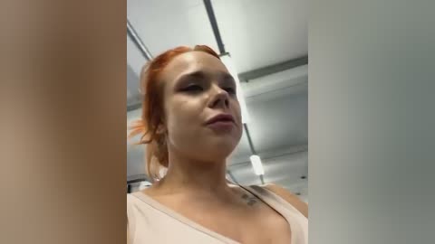 Media: Video of a fair-skinned woman with red hair, wearing a light-colored top, taken indoors in a modern, white-tiled room with visible ceiling lights.