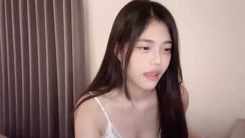 Media: Video of a young Asian woman with long black hair, fair skin, and small breasts, wearing a white spaghetti-strap top. She looks thoughtful, against a beige wall and vertical blinds.