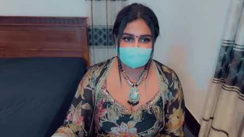 Media: Video of a South Asian woman with dark skin, wearing a colorful floral blouse, traditional jewelry, and a blue surgical mask, sitting on a bed with a wooden headboard.