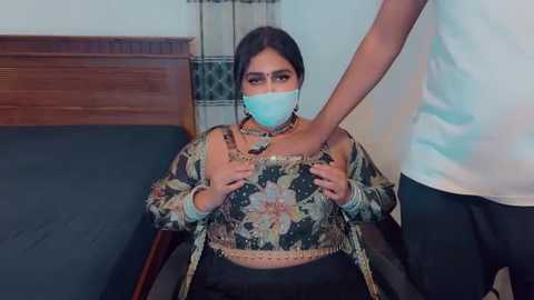 Media: Video of a woman in a floral top, blue mask, and silver jewelry, restrained by a man in a white shirt, in a dimly lit room.