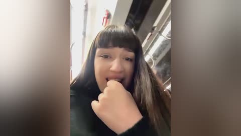 Media: A video of a young woman with long brown hair and bangs, biting her finger, wearing a black top, standing in a dimly lit room with metal pipes visible in the background.