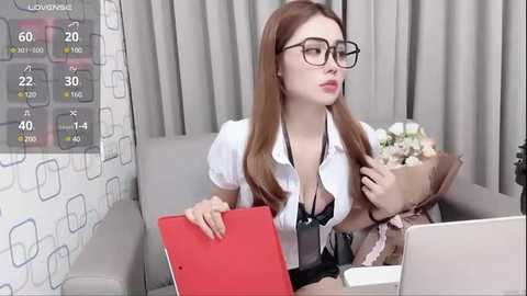 Media: A video shows a young woman with long brown hair, wearing glasses, a white blouse, and a black bra, sitting on a grey couch. She holds a red folder and a laptop.