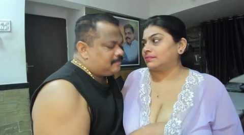 Media: Video of a middle-aged Indian man with a mustache in a black sleeveless shirt, standing next to a woman in a light purple, sheer, lace-trimmed robe. They are in an indoor setting with a framed picture of another man in the background.