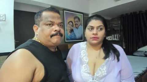 Media: Video of an overweight Indian man with a mustache, wearing a black sleeveless shirt, and a plus-size woman with dark hair in a ponytail, dressed in a lavender lace robe, standing in a bedroom with framed family photos on the wall.