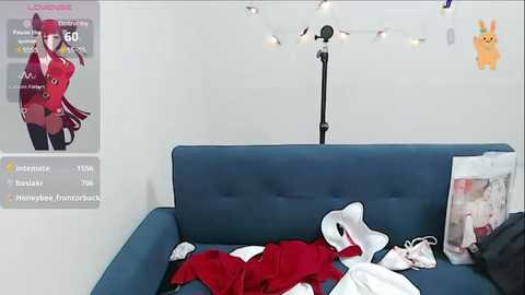 Media: Video of a modern, minimalist room with a blue couch, a white wall, and a black floor lamp. On the couch, a plush toy cat dressed in a red outfit lies next to a white handbag and a magazine.