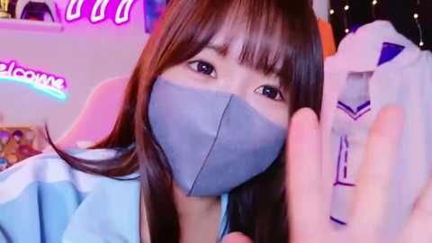 Media: Video of an East Asian woman with straight brown hair, wearing a light blue medical mask, light blue gown, and holding a white object with a purple emblem. Neon signs and colorful background.