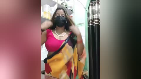 Media: Video of a woman in a vibrant sari with a black mask, adjusting her hair, in a domestic setting.