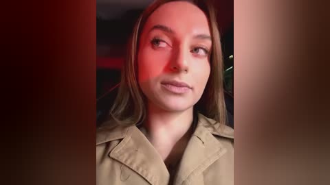 Media: Video of a young woman with fair skin, light brown hair, and brown eyes, wearing a tan trench coat, gazing slightly upward with a neutral expression. Background is blurred, with red and brown tones.