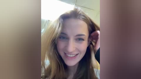 Media: Video of a smiling, fair-skinned woman with long, light brown hair, standing in a bathroom with beige tiles and a mirror, partially obscured by a person's arm.