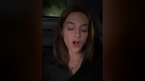 Media: A dimly-lit video captures a Caucasian woman with straight brown hair, wearing a black top, seated in a car, with her eyes closed and mouth slightly open.