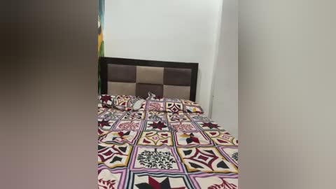 Media: Video of a neatly made bed with a colorful, geometric-patterned quilt and a dark wood headboard against a white wall. The room is simple and modern, with no visible furniture or decor.