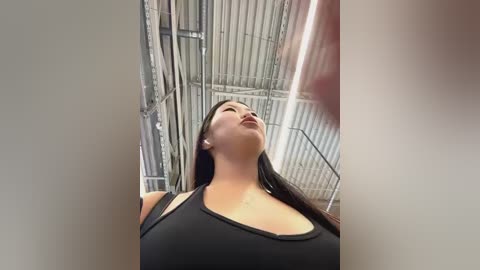 Media: Video of a woman with long black hair in a black tank top, standing in a tall, industrial-looking room with metal beams and a high ceiling.