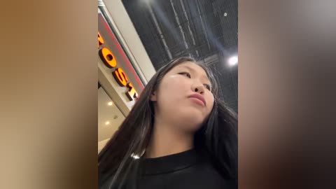 Media: A low-angle video of a young Asian woman with long black hair, wearing a black top, looking down, in a brightly lit indoor space with modern decor and a prominent \"Dunkin Donuts\" sign in the background.