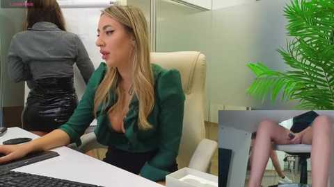 Media: A video of a blonde woman in a green blouse, leaning over a desk, with another woman in a grey shirt behind her. The setting is an office with a green plant in the background.