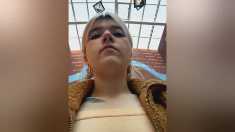 Media: A young woman with platinum blonde hair, wearing a brown leather jacket over a cream top, stands under a grid skylight. Her face is neutral, and the image has a blurred, slightly out-of-focus effect.