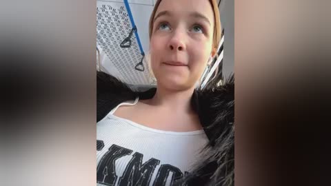 Media: Video of a young woman with light skin, light brown hair, and blue eyes, wearing a white tank top with black \"KIND\" text and black fur-trimmed jacket. She sits in a bus with a blue seat belt and mesh seat.